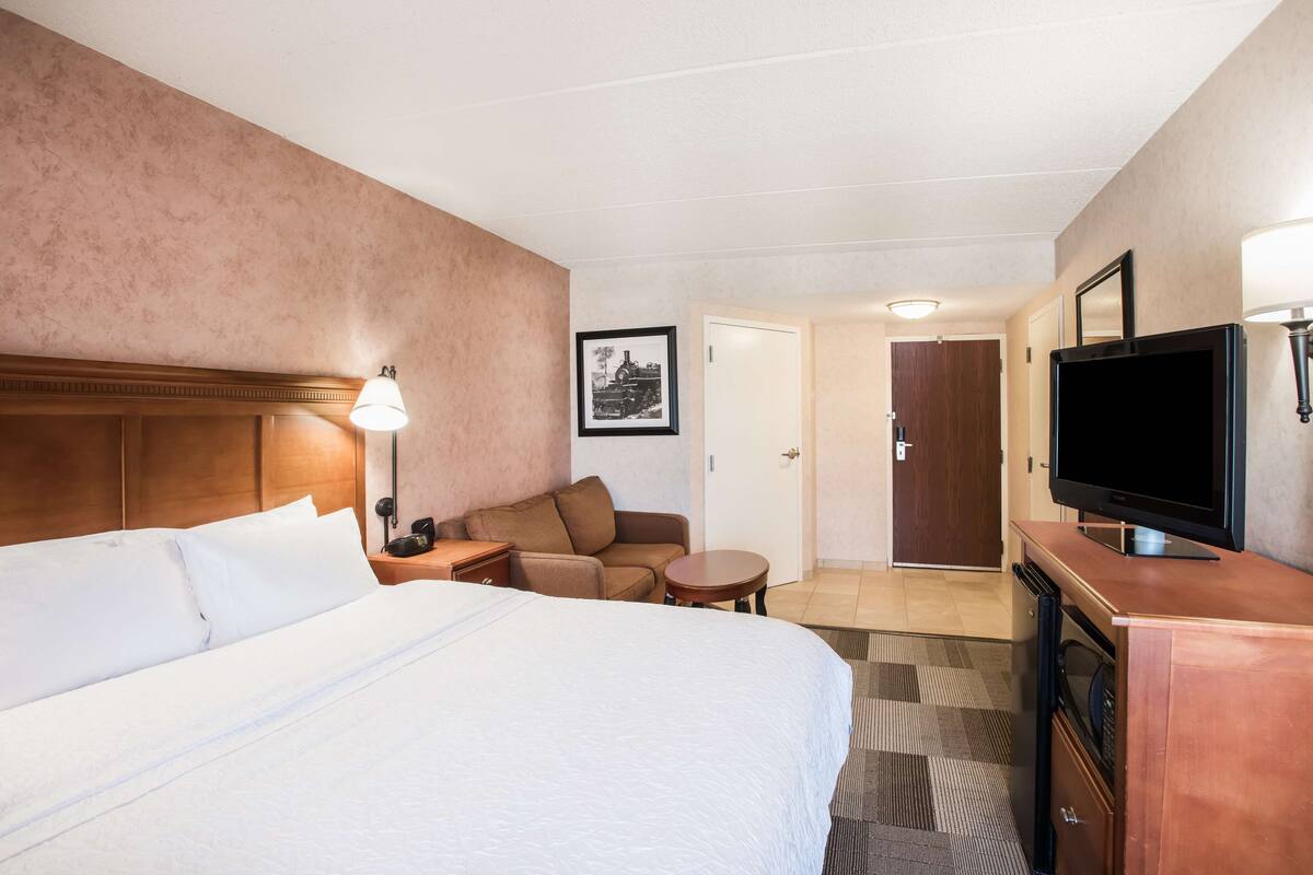 A hotel room with a large bed, a sofa, a small table, a TV, and a door leading to another room.