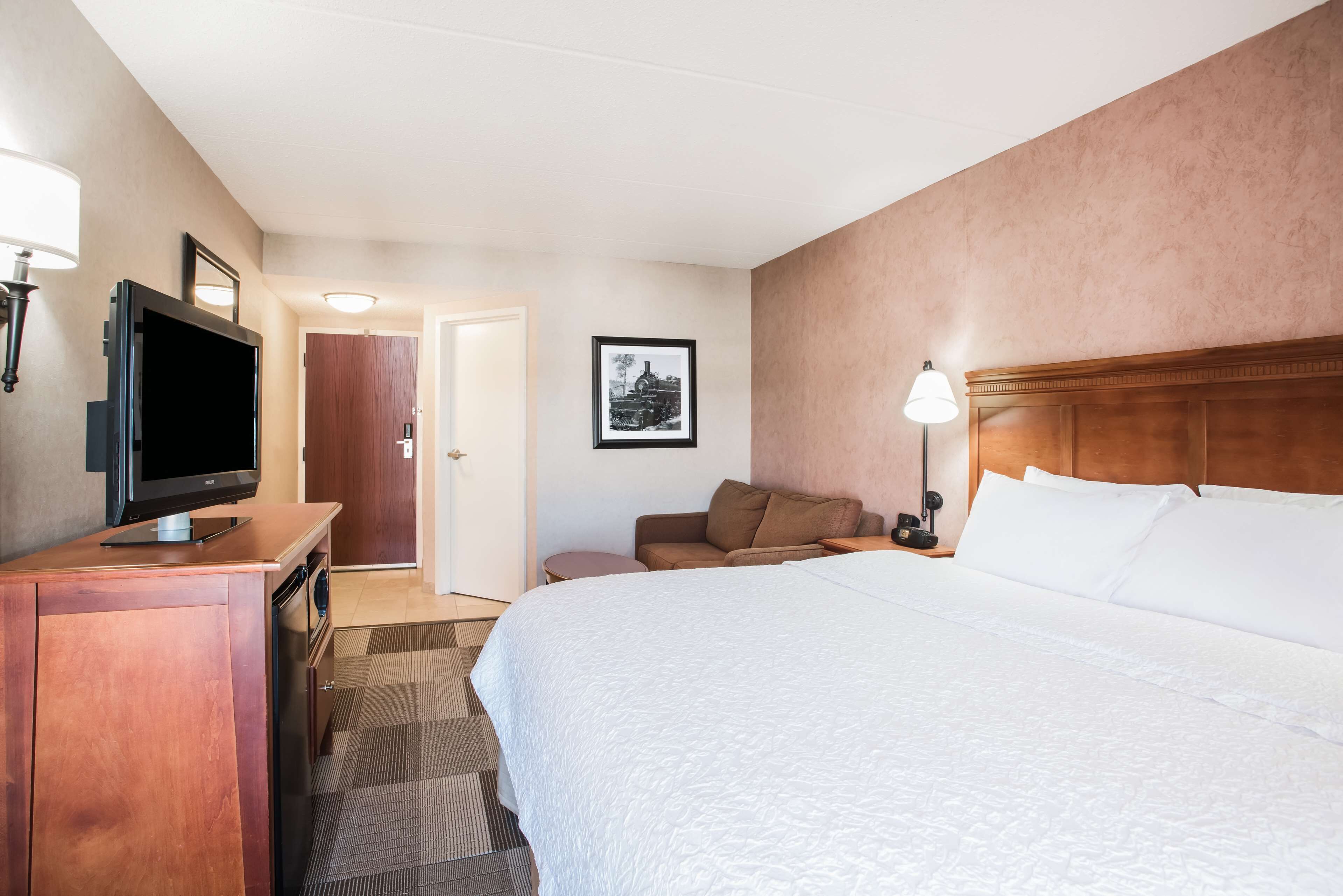 A hotel room with a bed, a desk with a television, a nightstand with a lamp, a framed picture on the wall, and a door leading to another room.