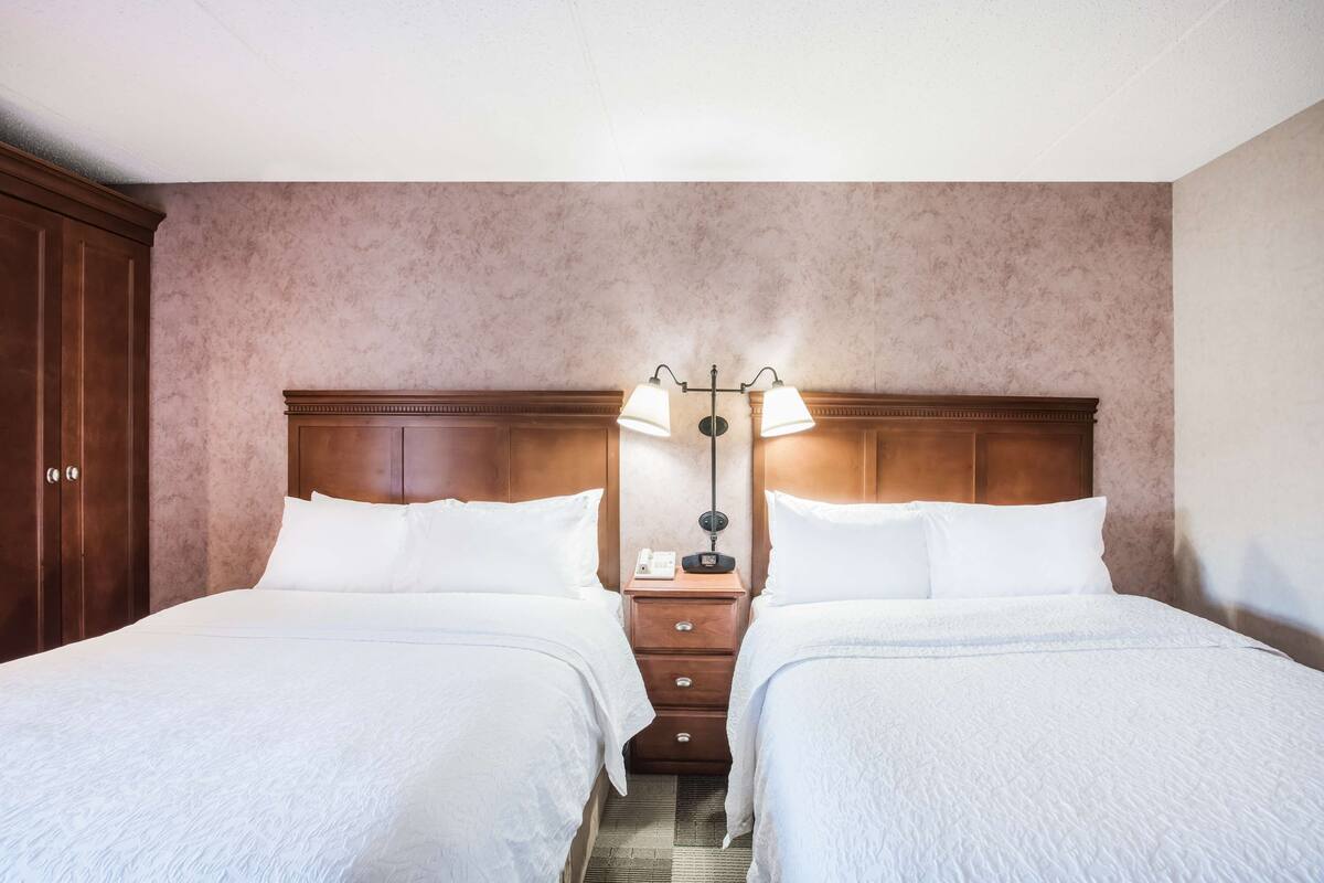 A hotel room with two beds, a wooden headboard, a nightstand with a lamp, and a telephone.