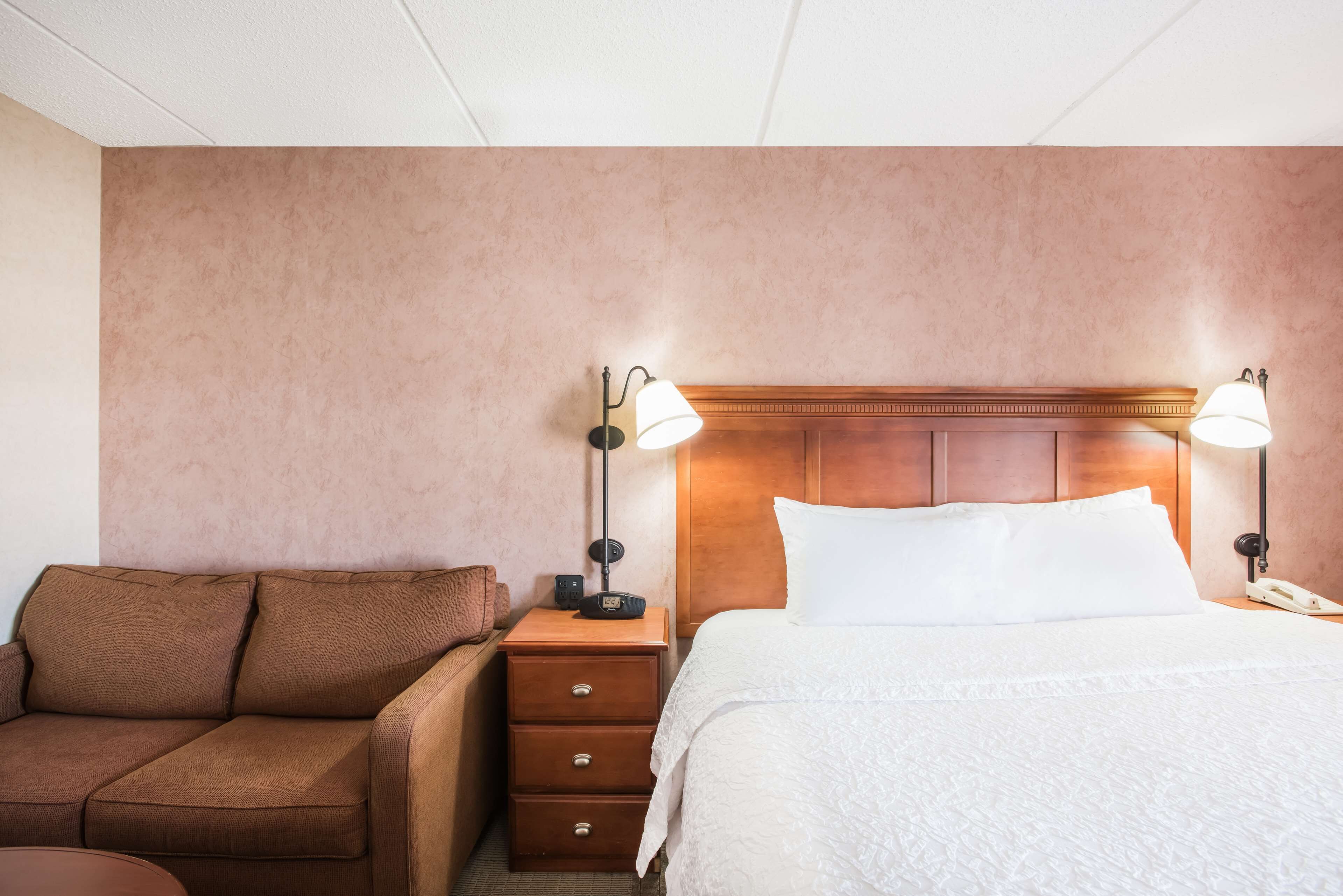 A hotel room with a brown sofa, a wooden nightstand with a lamp, a bed with white linens, and a wooden headboard.