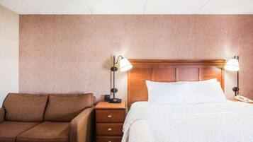 A hotel room with a brown sofa, a wooden nightstand with a lamp, a bed with white linens, and a wooden headboard.
