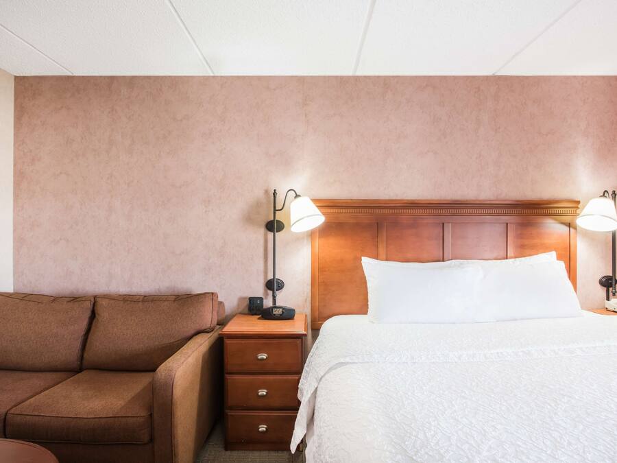 A hotel room with a brown sofa, a wooden nightstand with a lamp, a bed with white linens, and a wooden headboard.