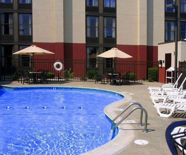 A swimming pool with lounge chairs and umbrellas, surrounded by a fence and a building with large windows.