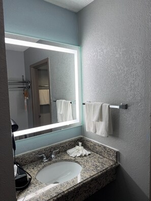 Un baño con encimera de granito, un lavabo blanco, un espejo grande con luz y un perchero con dos toallas.
