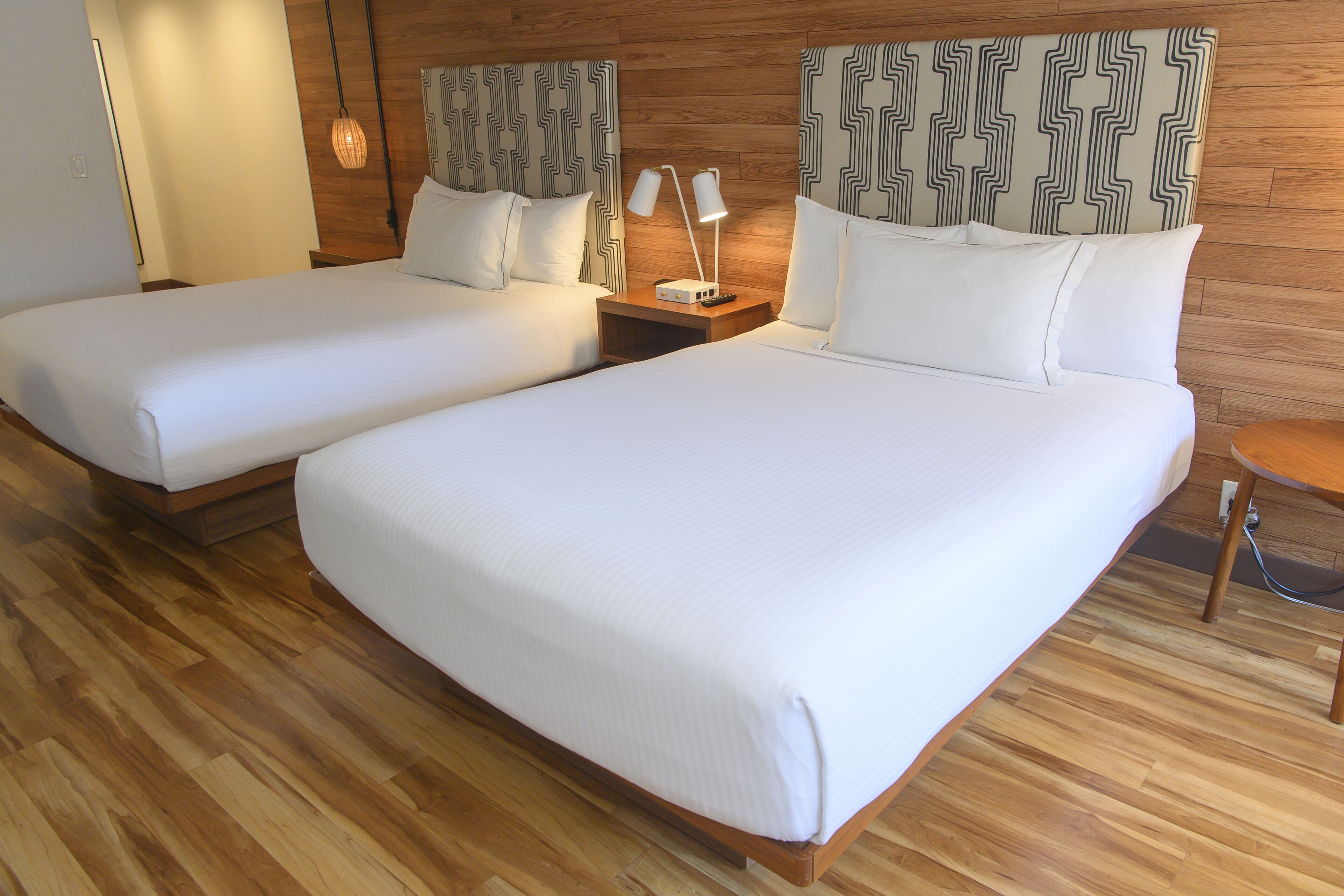 A hotel room with two single beds, wooden flooring, a nightstand with a lamp, and patterned headboards.