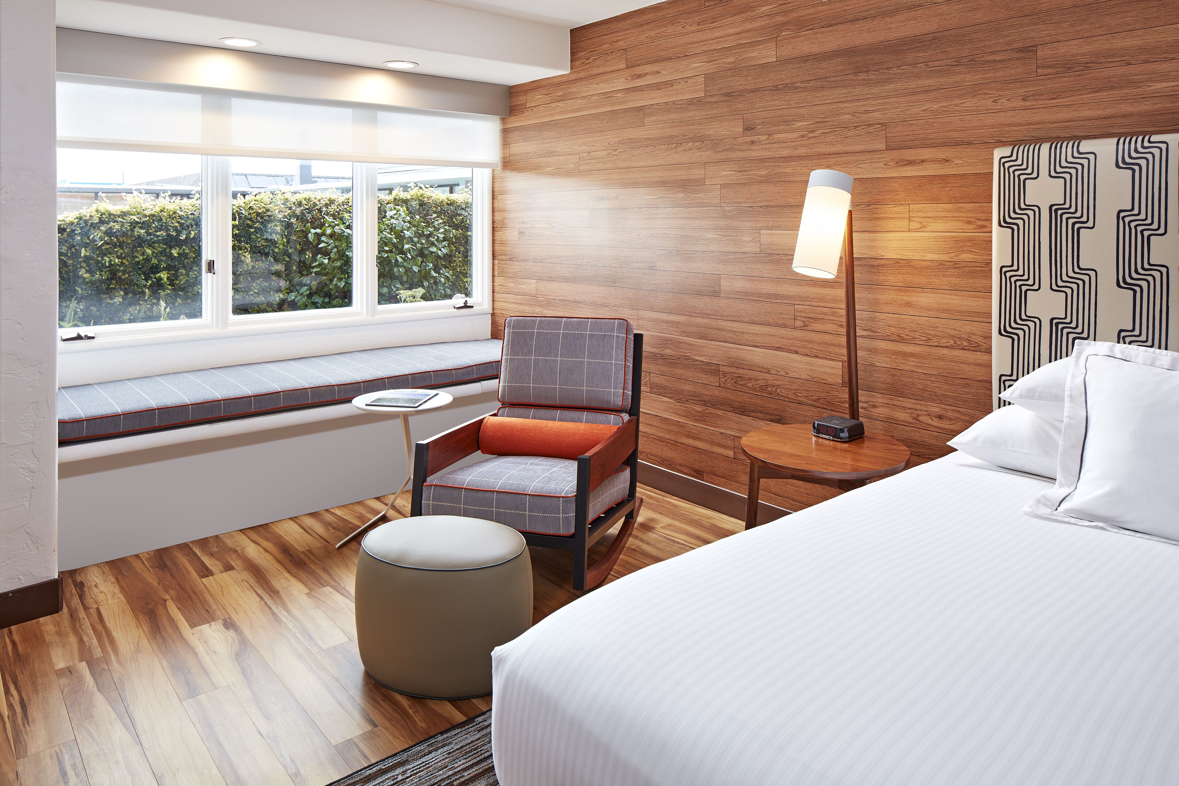 A modern hotel room with a large bed, a seating area with a chair and ottoman, a bedside table, and a view of greenery through the window.
