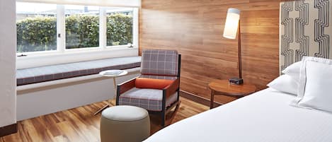 A modern hotel room with a large bed, a seating area with a chair and ottoman, a bedside table, and a view of greenery through the window.