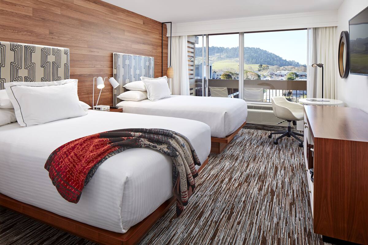 A hotel room with two beds, a desk, and a view of the countryside.