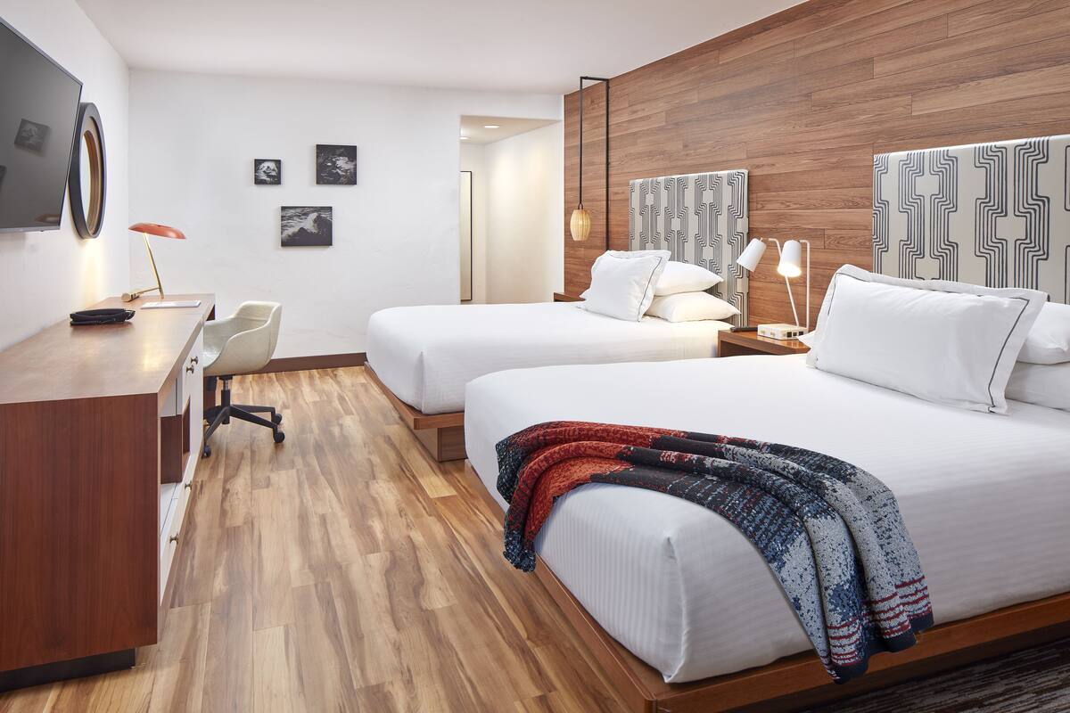A hotel room with two beds, a desk, a chair, and a TV.