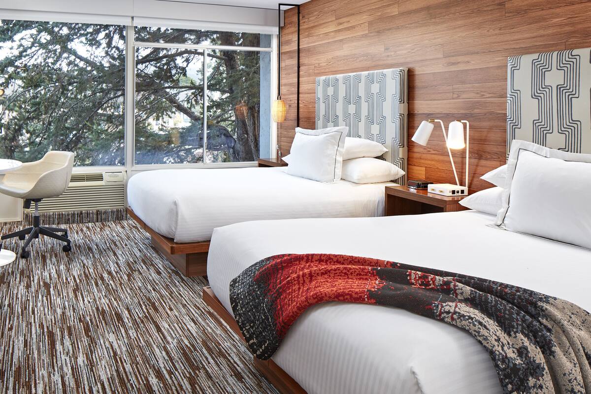 A hotel room with two beds, a desk, a chair, and a large window offering a view of trees.