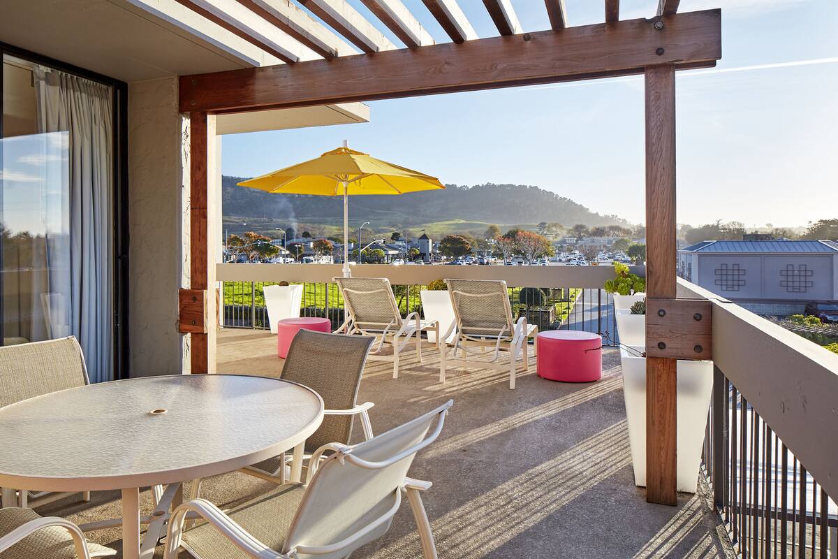 A balcony with a table and chairs, a yellow umbrella, and a view of a parking lot and hills.