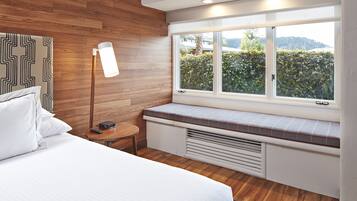 A bedroom with a wooden headboard, a bedside table with a lamp, a window seat with a view of greenery, and a bed with white linens.