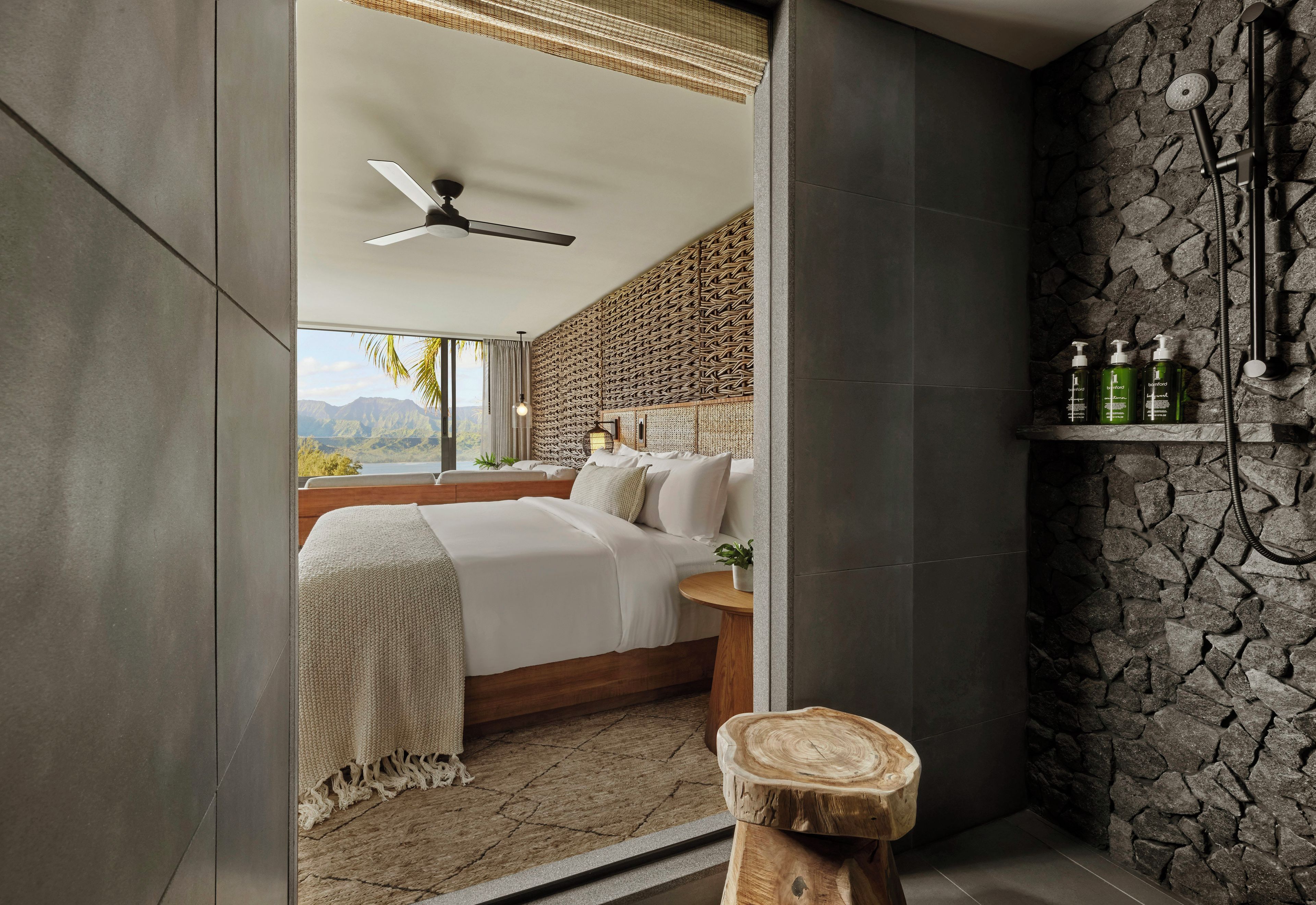 Un dormitorio moderno con una cama grande, ventilador de techo, vista a montañas y pared de piedra con artículos de tocador.