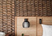 A room with a woven wall, a bed with white bedding, a round side table with a geometric pattern, and a black hanging light fixture.