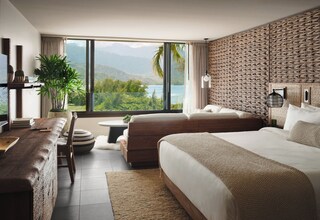 A modern hotel room with a large bed, a sofa, a dining area, and a view of mountains and water.