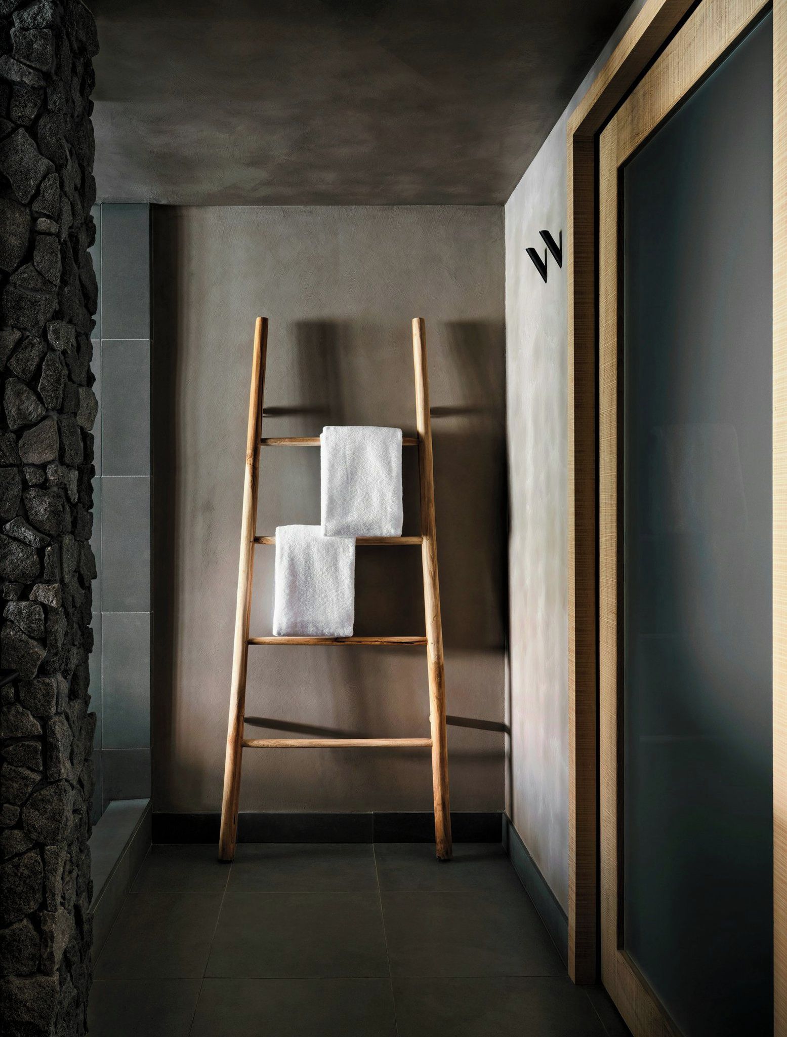 A wooden ladder with two white towels against a wall with a stone texture on one side and a dark door on the other.