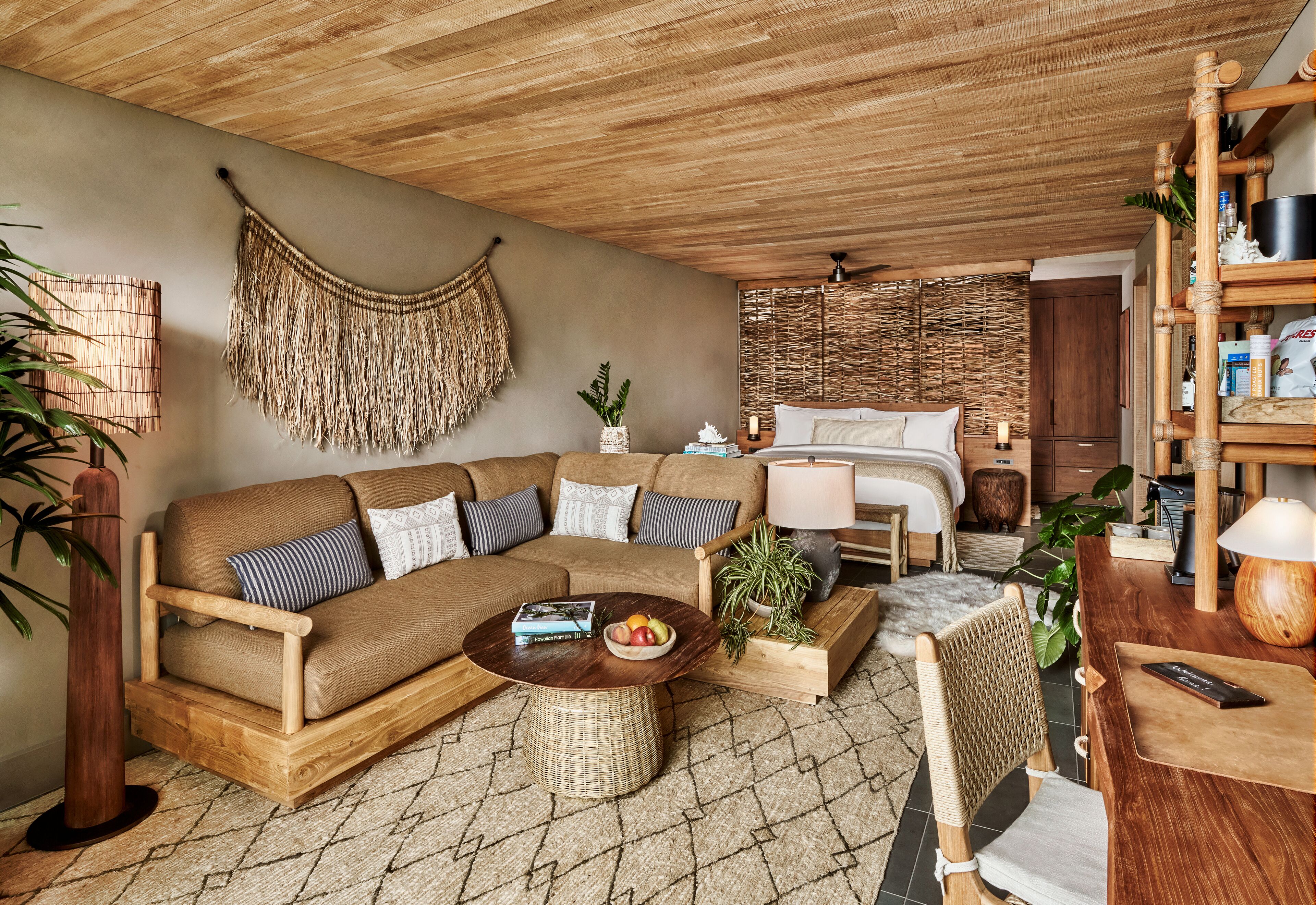 A cozy living room with a wooden sofa, a round coffee table with fruits, a wicker chair, and a bed with a woven headboard.