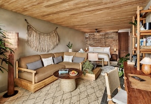 A cozy living room with a wooden sofa, a round coffee table with fruits, a wicker chair, and a bed with a woven headboard.