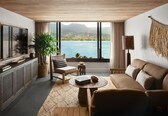 A living room with a large window offering a view of a lake and mountains, a sofa, a chair, a coffee table, and a TV on a wooden cabinet.