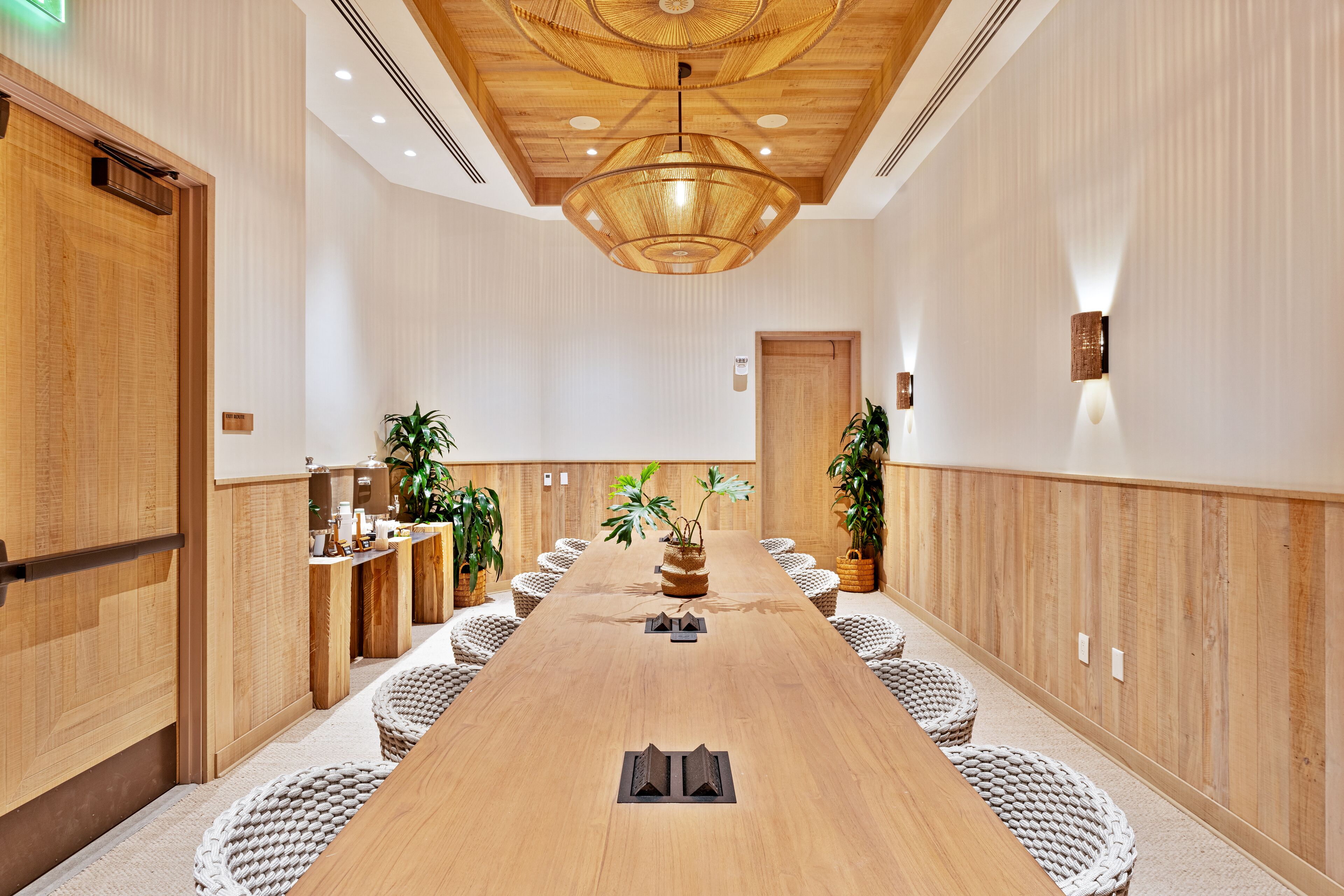 A conference room with a long wooden table, white woven chairs, and a central plant.