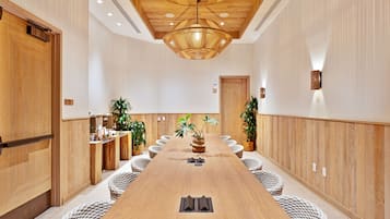 A conference room with a long wooden table, white woven chairs, and a central plant.