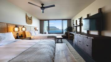 A hotel room with two beds, a large window with a view of the sea, a ceiling fan, and a flat-screen TV mounted on the wall.