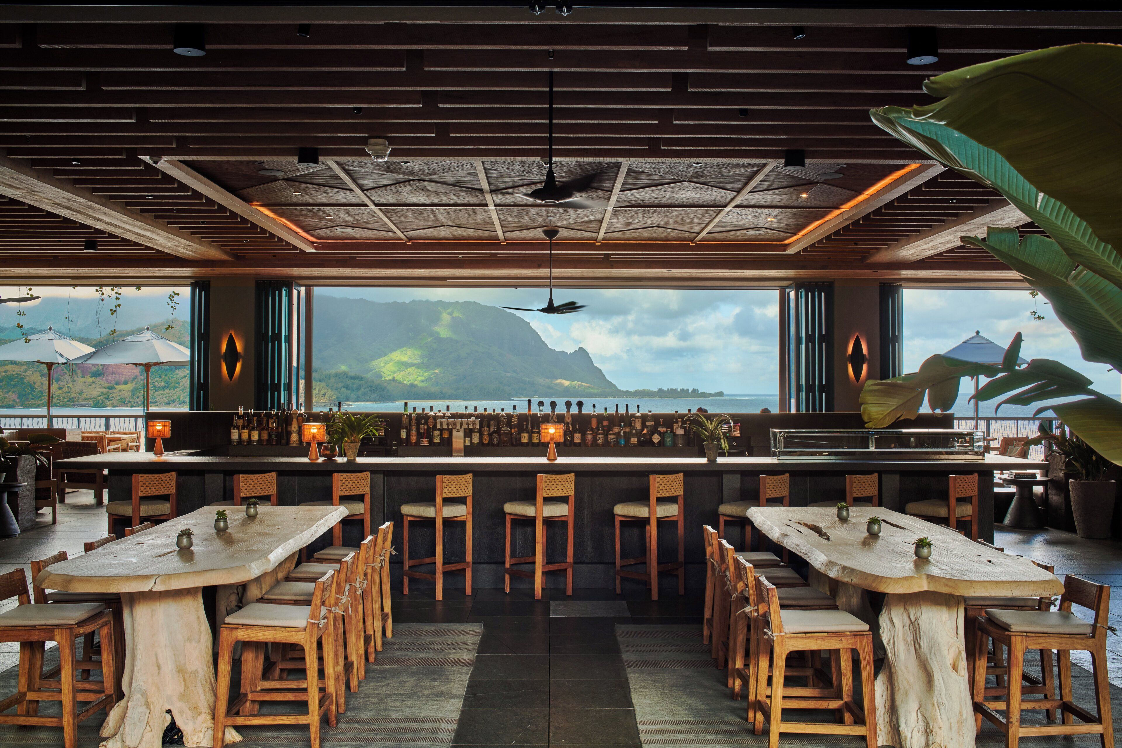 Um restaurante com uma longa mesa de madeira e cadeiras, área de bar e grandes janelas com vista para uma montanha e oceano.