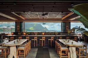 A restaurant with a long wooden table and chairs, a bar area, and large windows offering a view of a mountain and ocean. - 1 Hotel Hanalei Bay (Princeville)