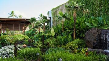 A lush garden with a waterfall, dense foliage, and a modern building with a sloped roof.