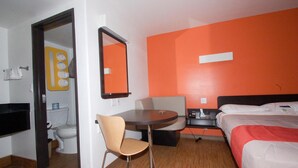 A hotel room with a bed, a desk, a chair, a bathroom with a toilet and sink, and an orange accent wall. - Motel 6 Victorville - Desert Express (Victorville)