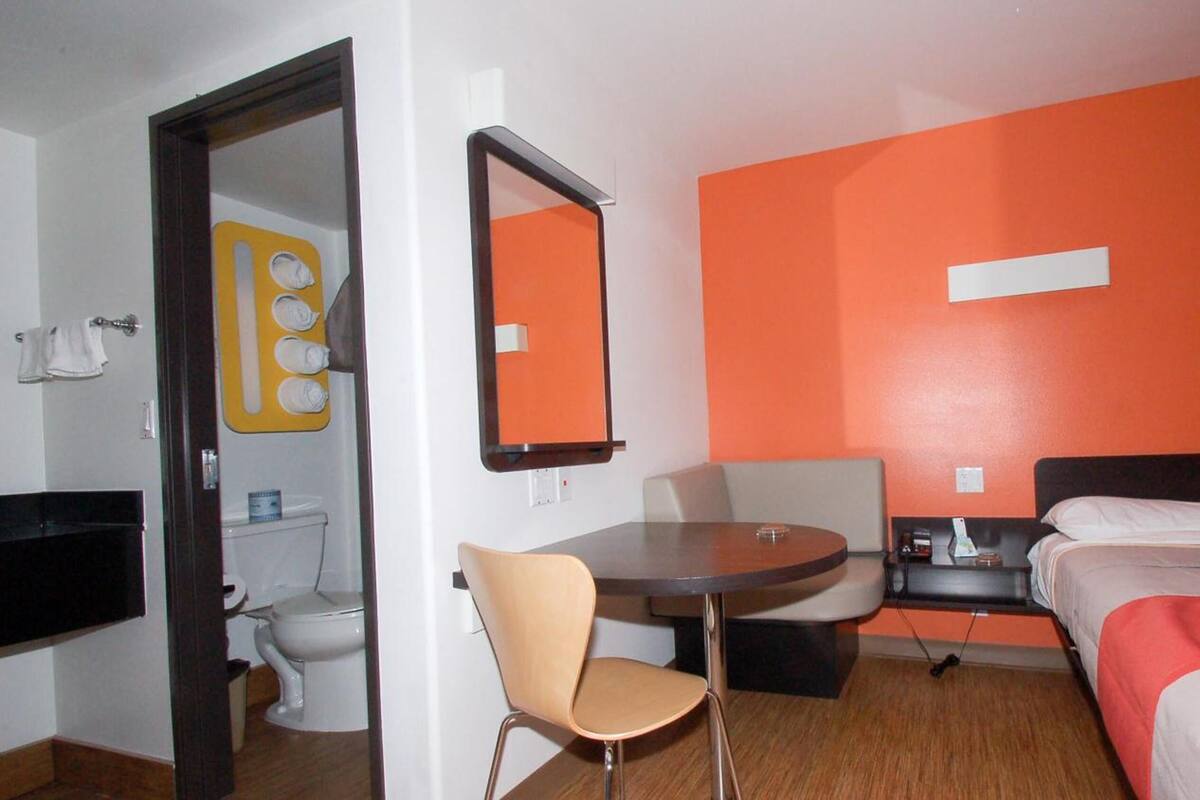 Une chambre d’hôtel comprenant un lit, un bureau, une chaise, une salle de bain avec des toilettes et un lavabo, ainsi qu’un mur d’accent de couleur orange.