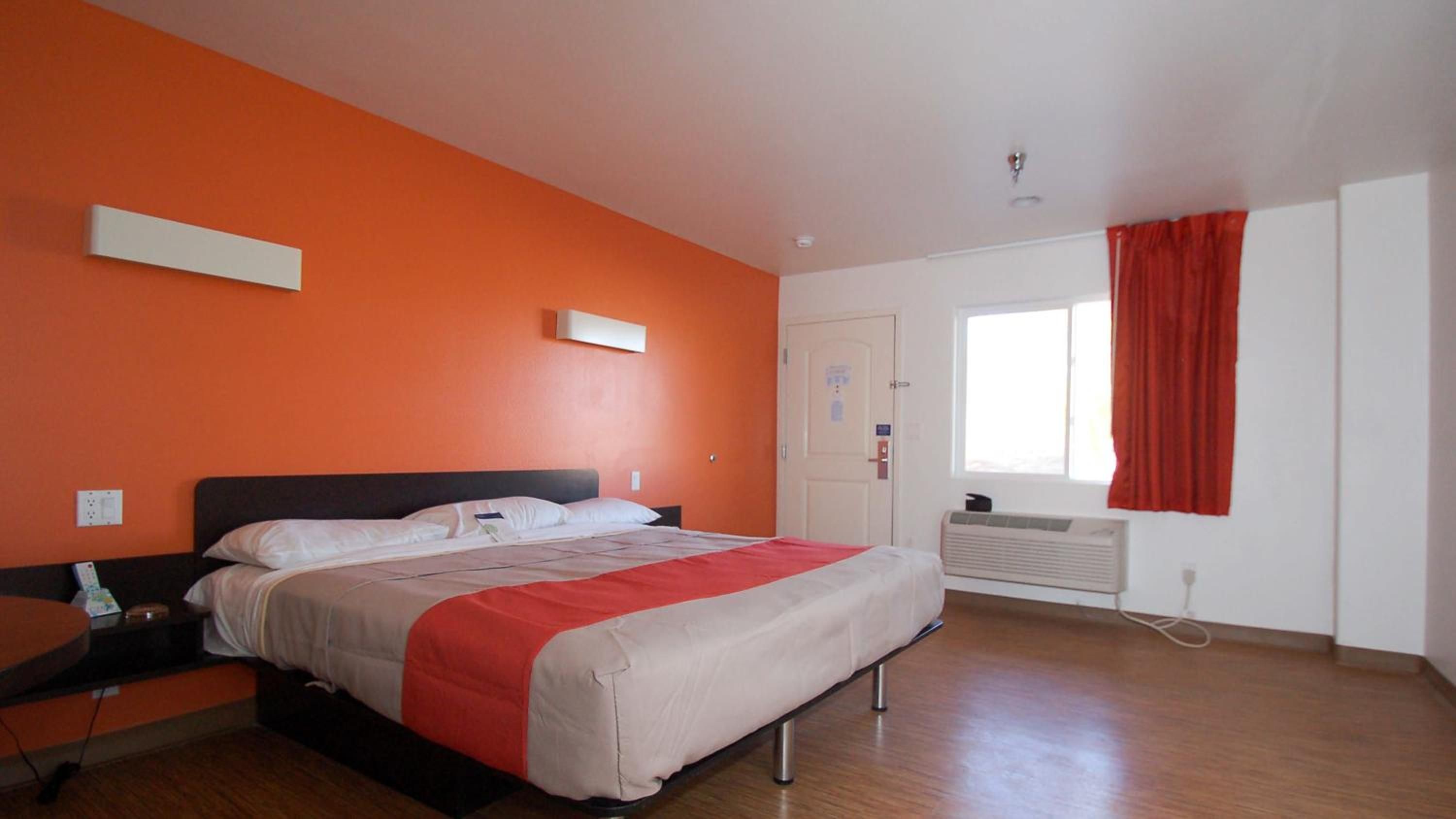 Habitación de hotel con cama, mesita de noche, ventana con cortinas rojas y un calefactor en la pared.