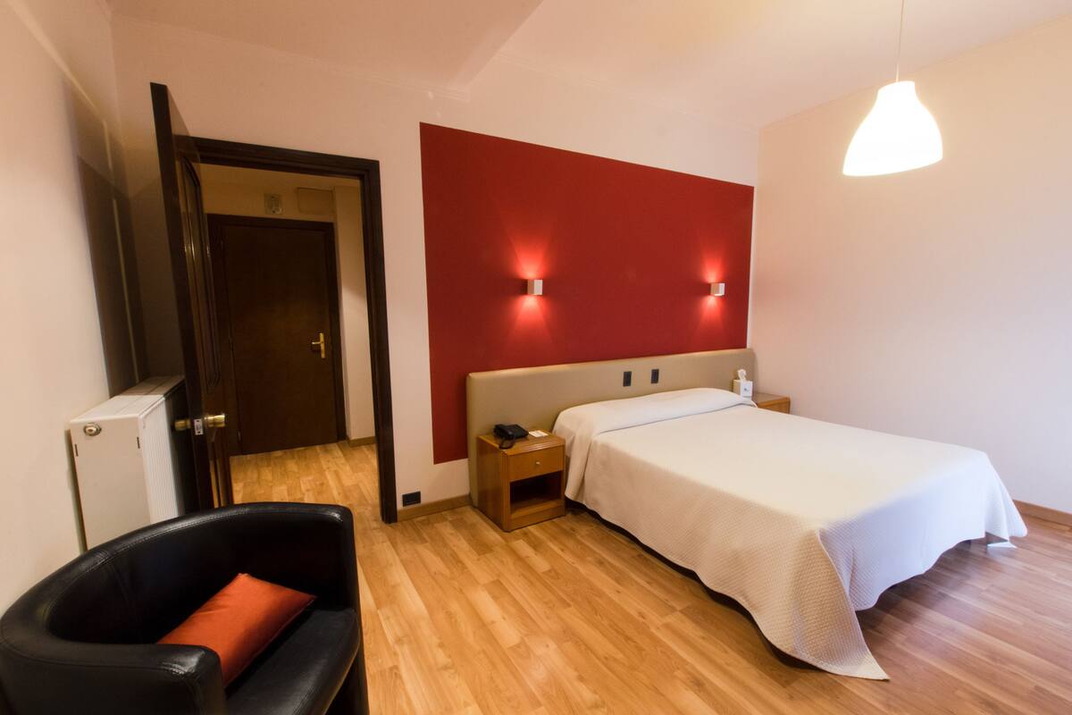 A hotel room with a wooden floor, a bed with a white bedspread, a nightstand with a phone, a red accent wall, and a black chair with an orange cushion.