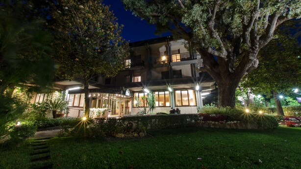 Un hôtel à plusieurs étages, doté d’une entrée bien éclairée, entouré d’une végétation luxuriante et d’un grand arbre.