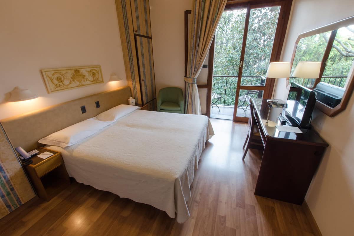 A hotel room with a bed, a desk with a computer, a chair, a small table, and a window with curtains.