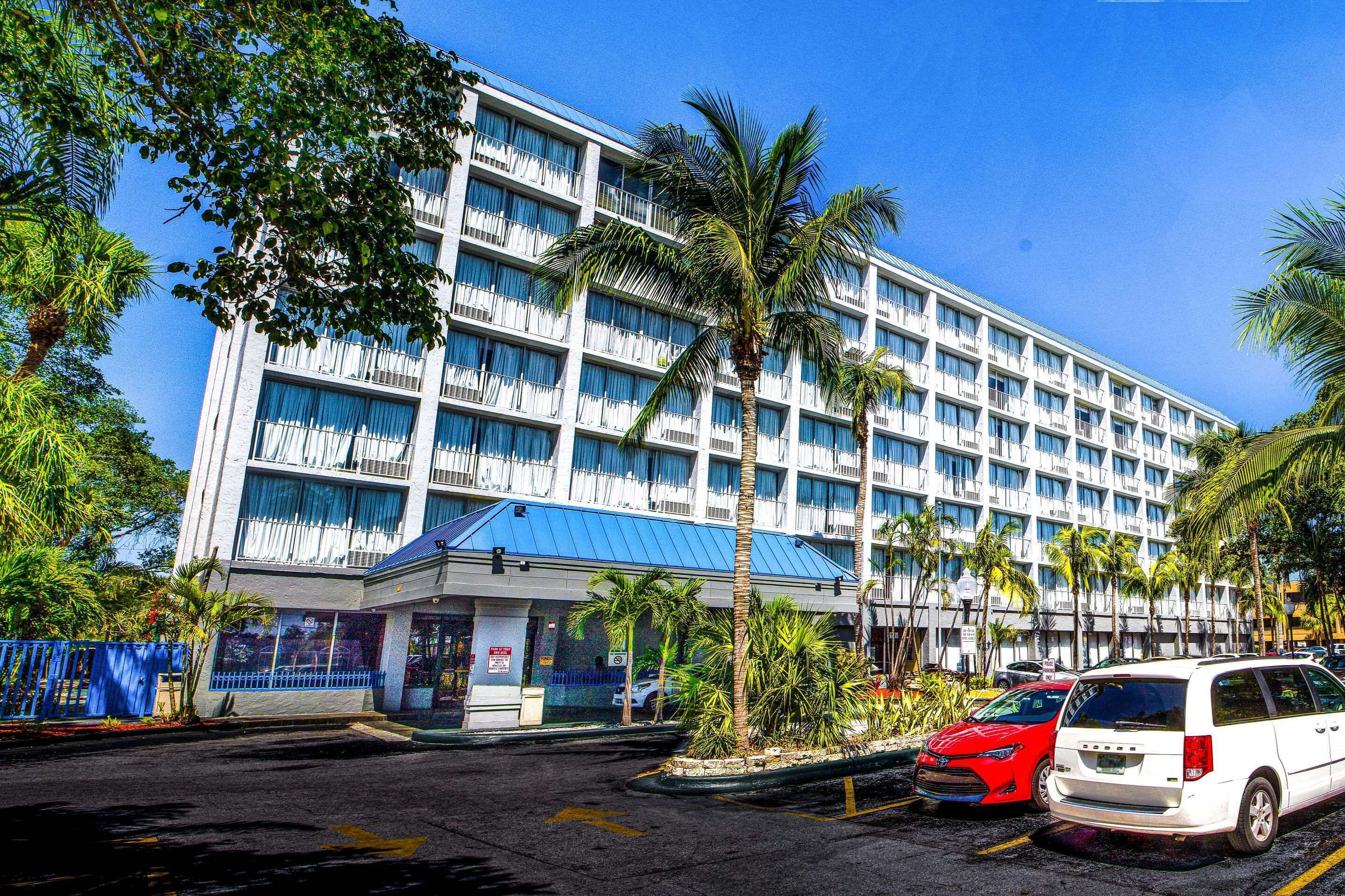 Un hotel de varios pisos con techo azul, palmeras y un estacionamiento con varios automóviles.