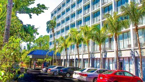 A multi-story hotel with a row of cars parked in front, surrounded by palm trees.
