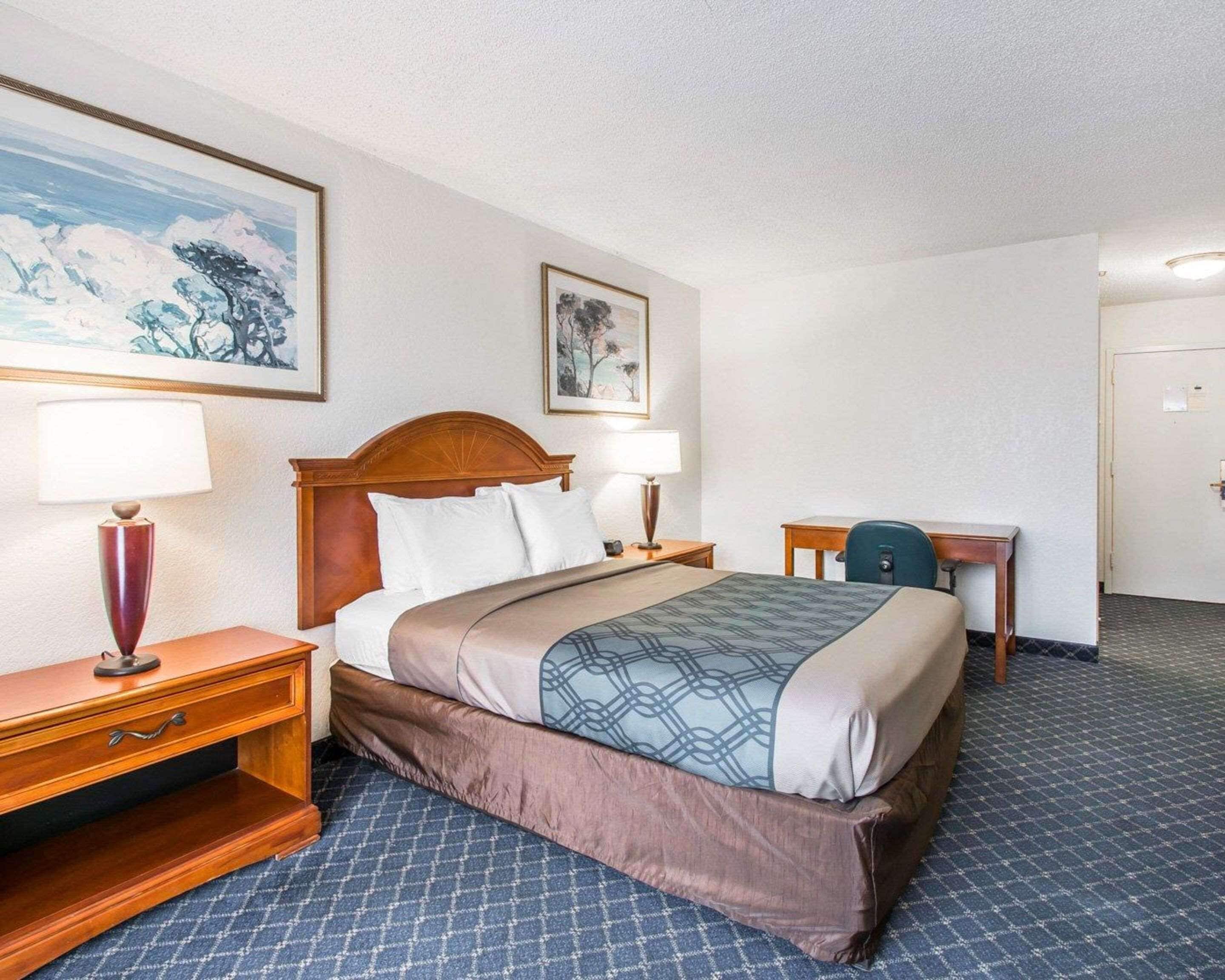 Habitación de hotel con una cama grande, dos mesitas de noche con lámparas, un escritorio y dos cuadros en la pared.