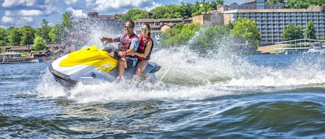 Una pareja en un jet ski creando olas en el agua, con un hotel y edificios al fondo.