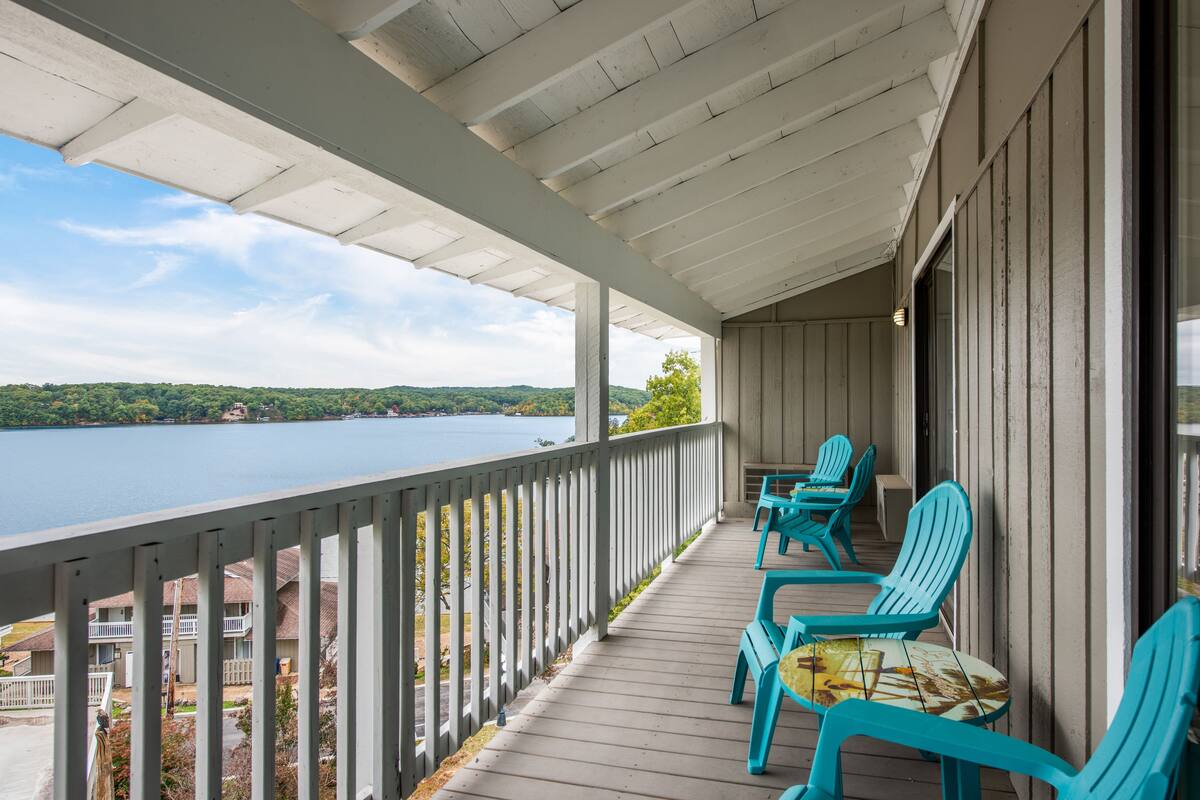 Ein überdachter Balkon mit Blick auf einen See, mit türkis-farbenen Stühlen und einem Tisch.