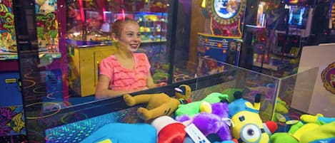 Une femme est assise dans une salle d’arcade bien éclairée, avec en arrière-plan divers jouets en peluche colorés et des machines d’arcade.