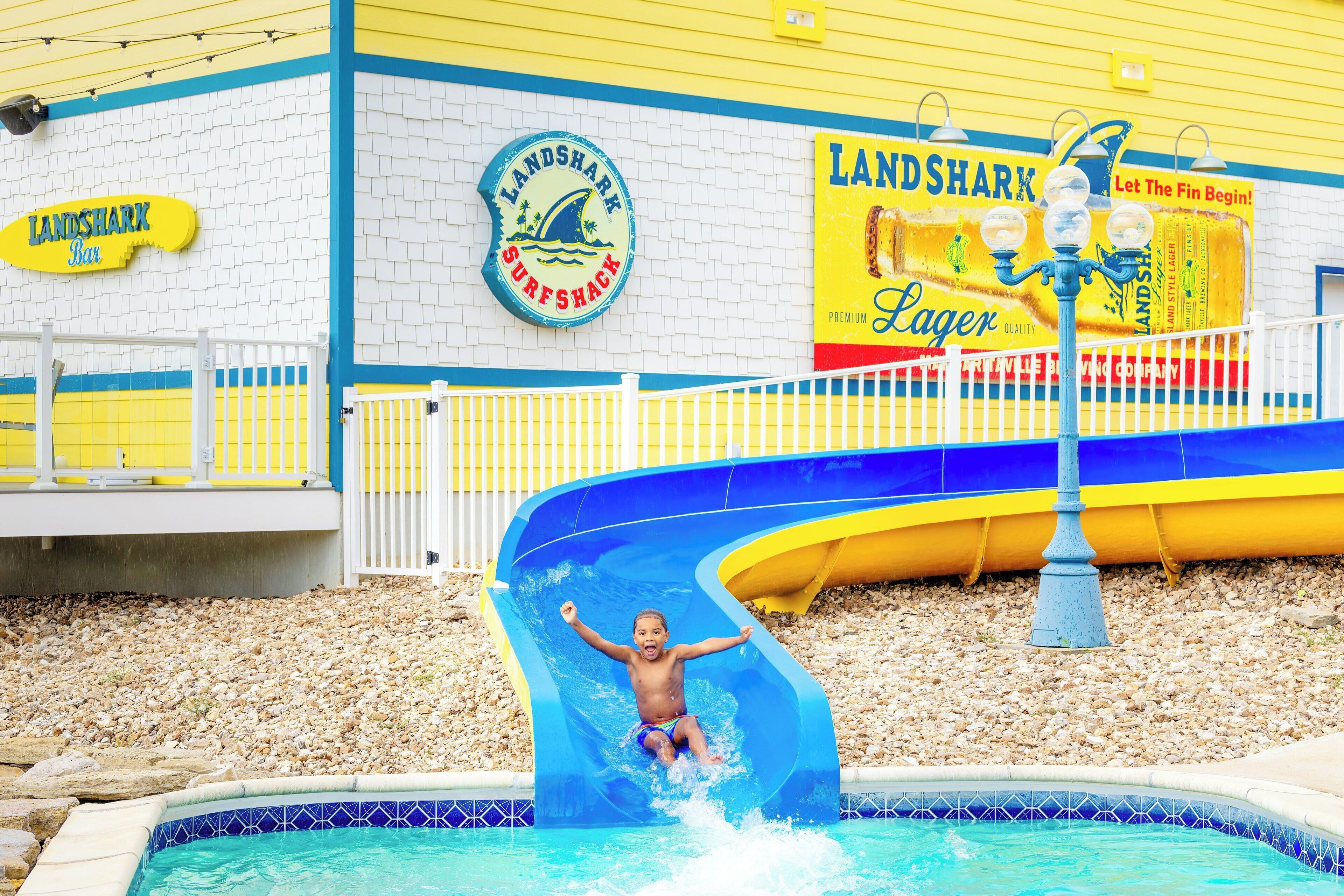 Niño deslizándose en una piscina dentro de un parque acuático, con un edificio de temática surfista al fondo.