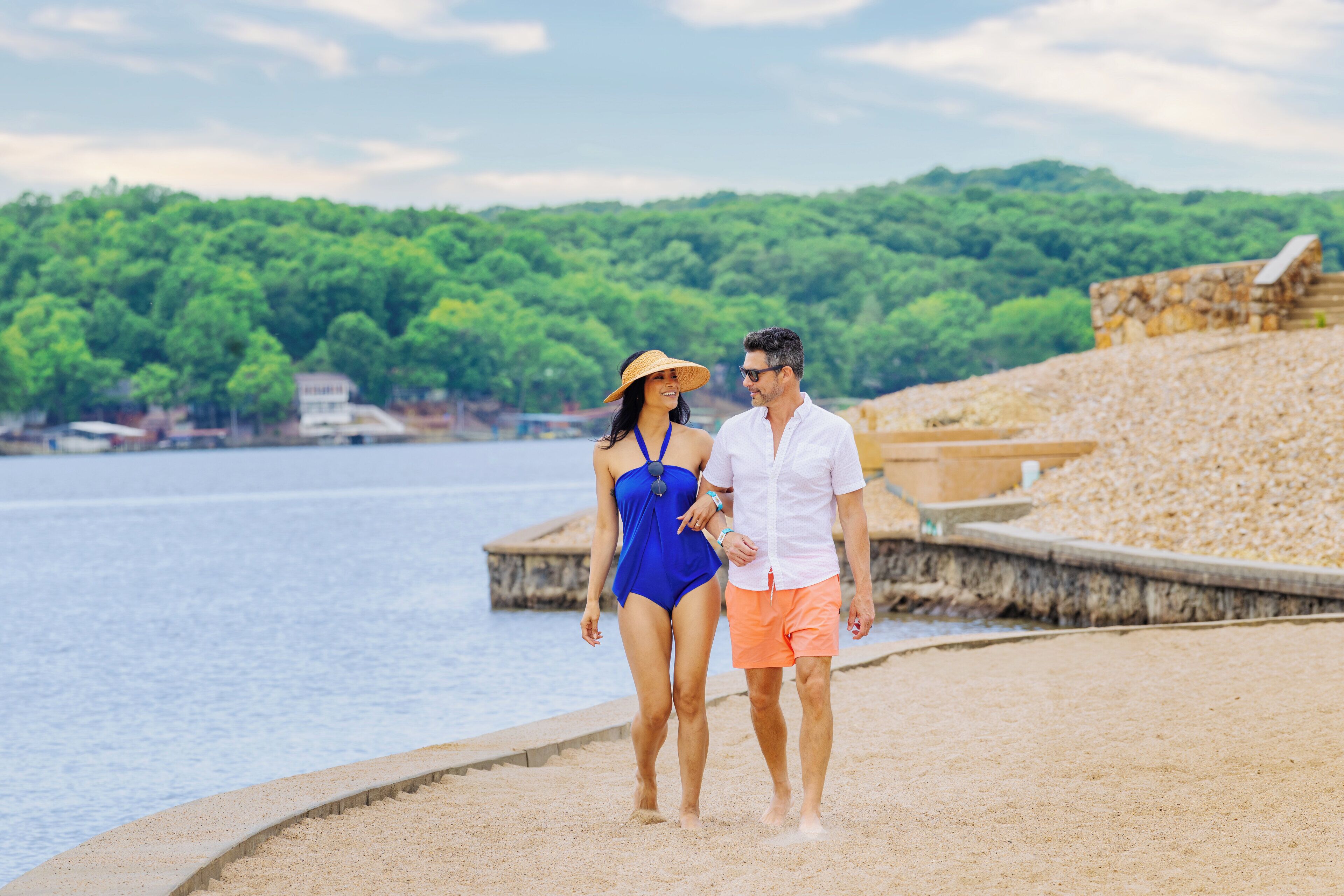 A couple walking on a sandy beach with a stone wall and greenery in the background.