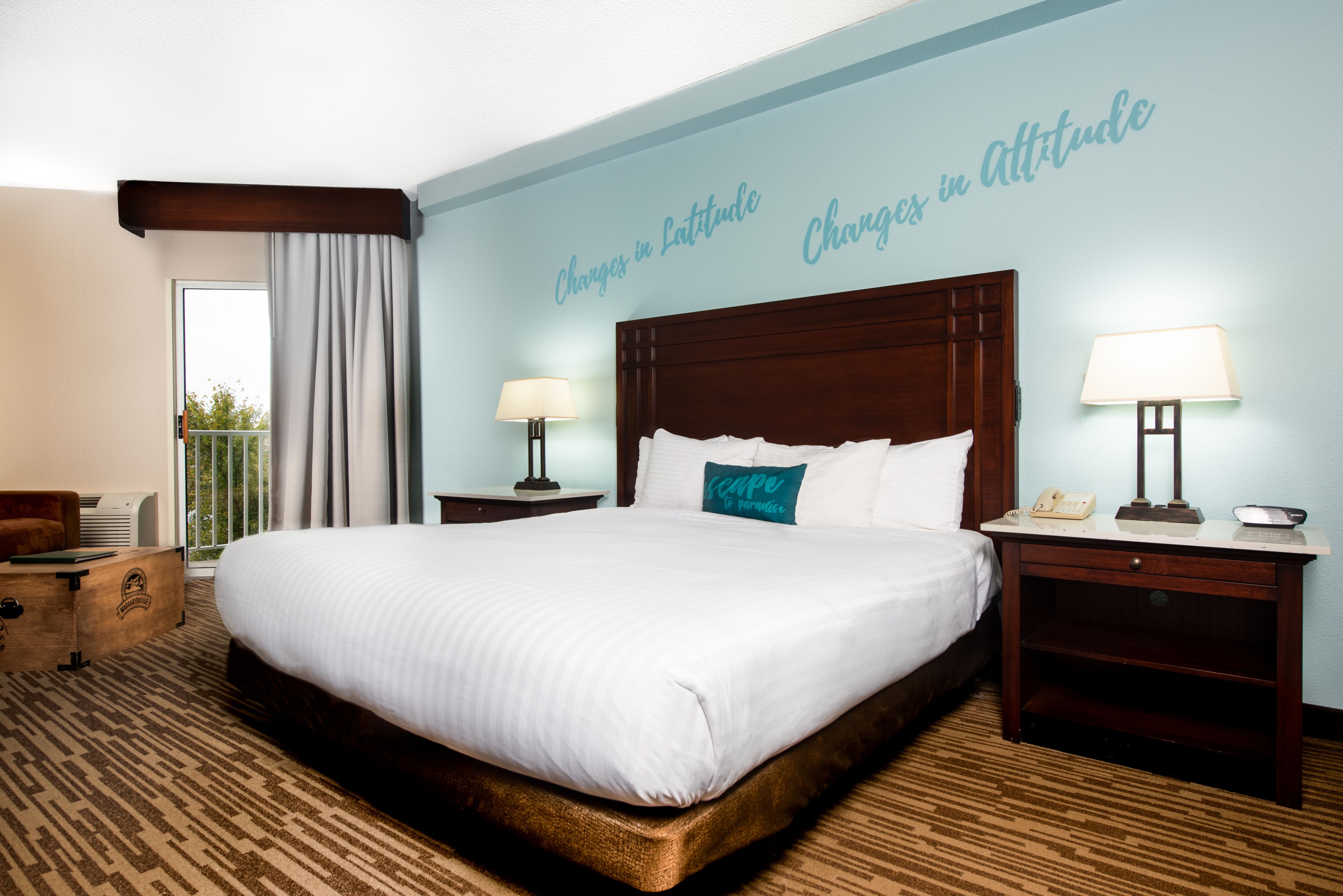 A hotel room with a large bed, two bedside tables with lamps, a wooden nightstand, a chair, and a view of the outdoors.