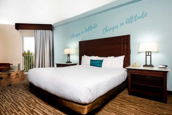 Habitación de hotel con una cama grande, dos mesitas de noche con lámparas, una mesita auxiliar de madera, una silla y vistas al exterior.