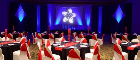 A banquet hall with round tables, white chairs with red sashes, and a stage with a floral projection.