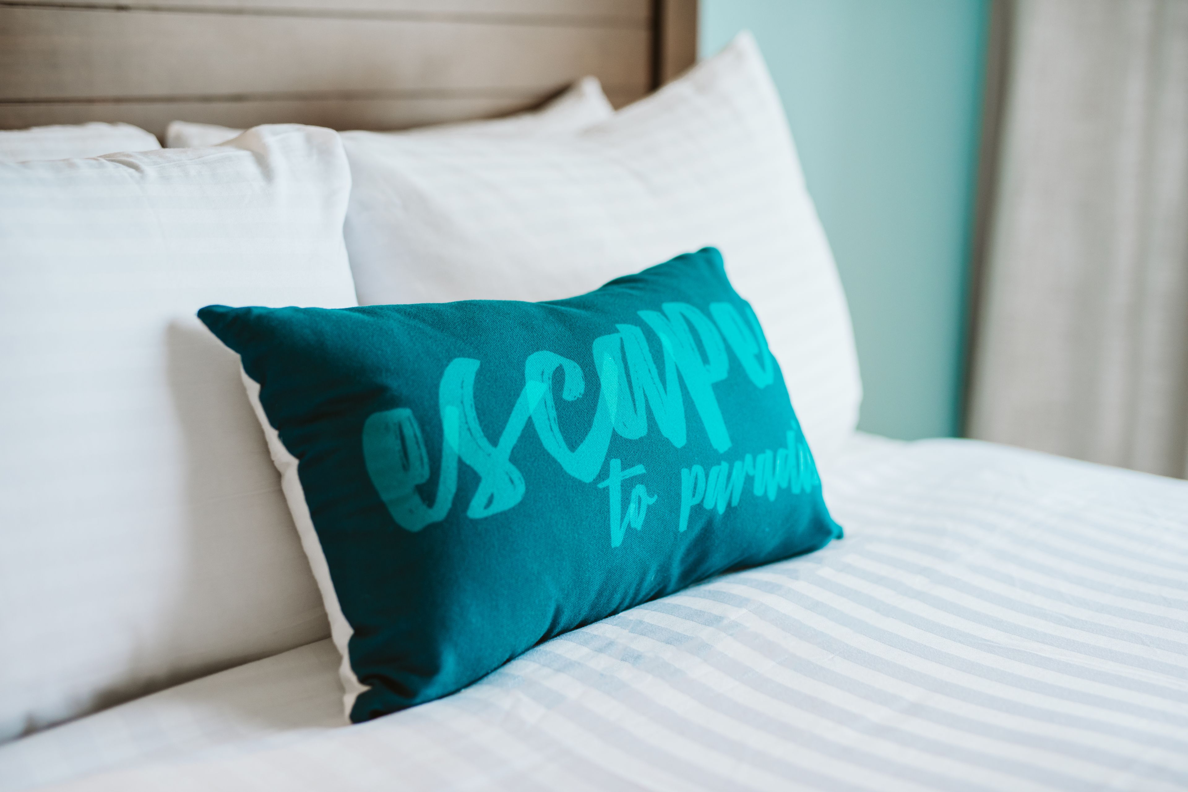 A teal pillow with the text "escape to paradise" on a bed with white bedding and a wooden headboard.