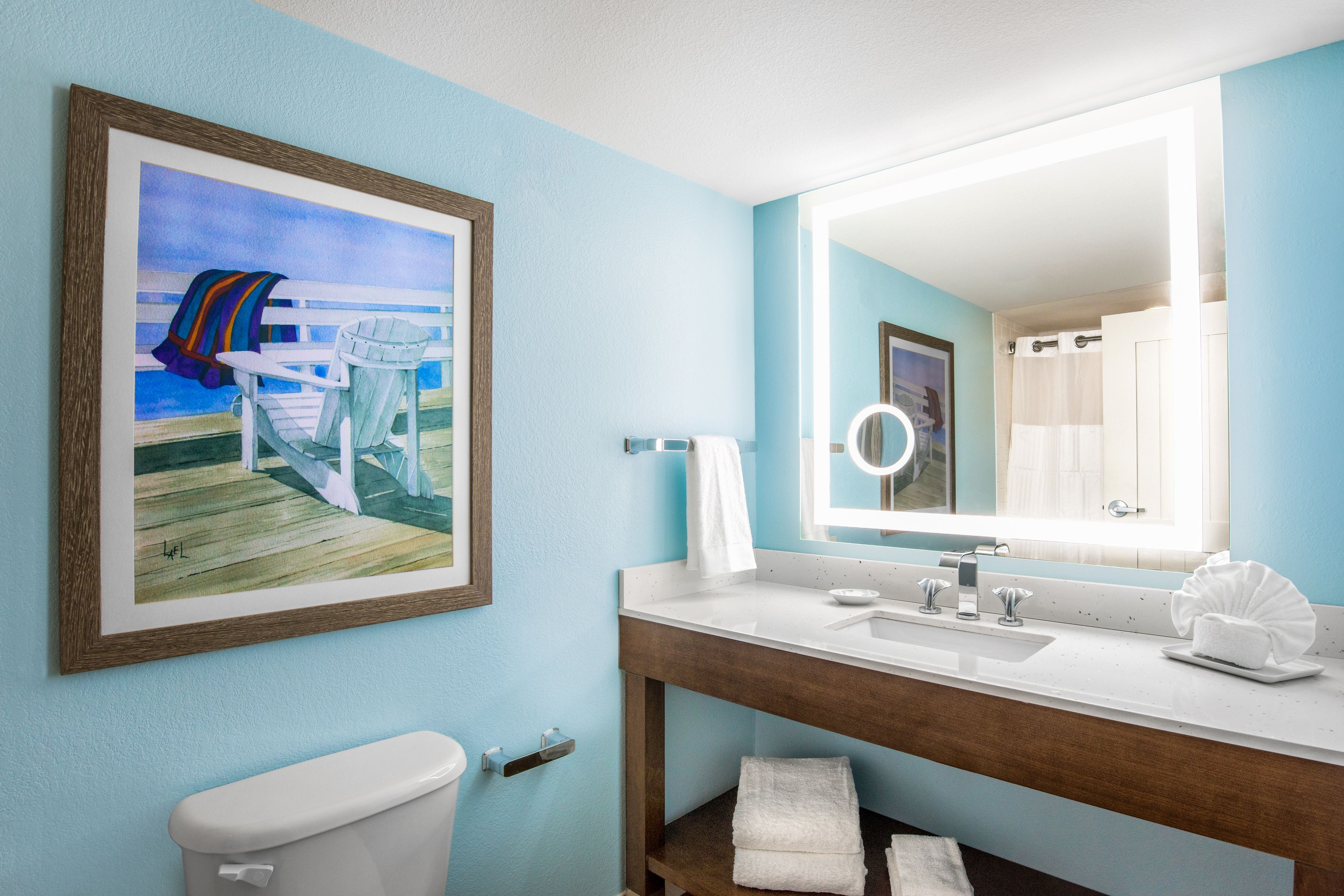 A bathroom with a painting of a beach scene, a wooden vanity, a white sink, and a mirror with a light.