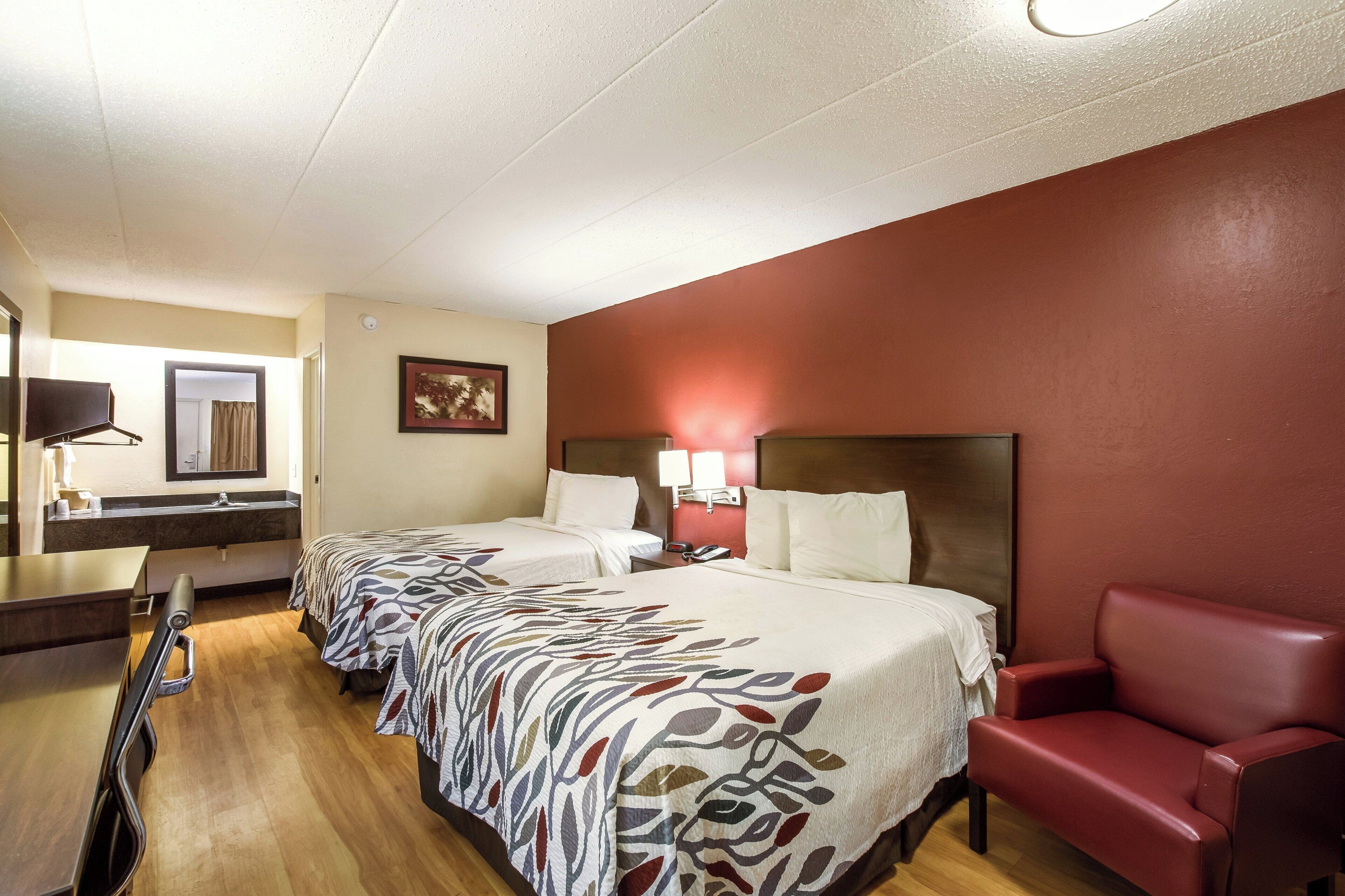 Een hotelkamer met twee bedden, een rode fauteuil, een bureau met spiegel en een schilderij aan de muur.