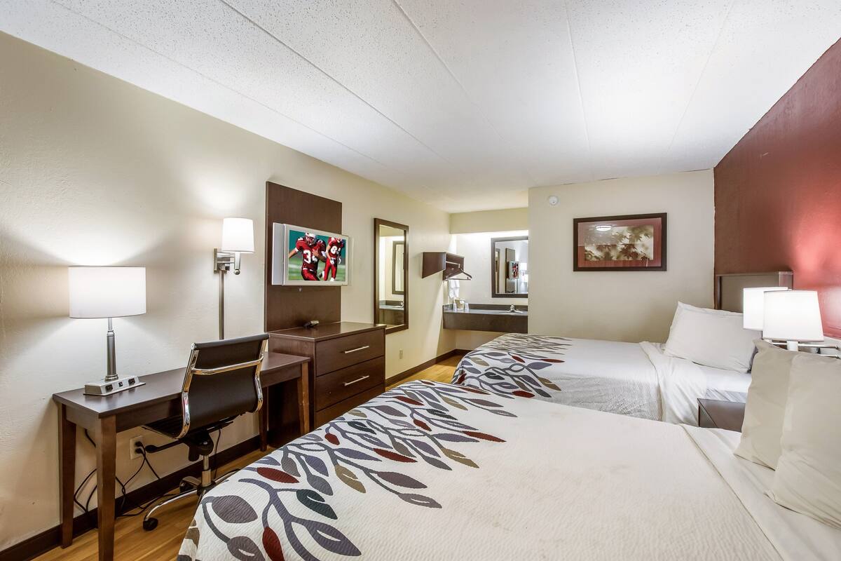 A hotel room with two beds, a desk with a chair, a dresser, a mirror, and a painting on the wall.